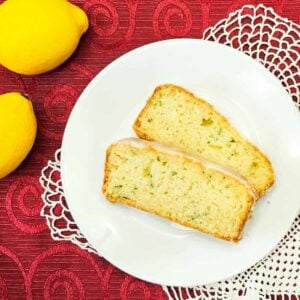 lemon zucchini bread sliced on a white plate sitting on a red table cloth with lemons to the side of it with a doily under the plate
