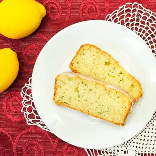 lemon zucchini bread sliced on a white plate sitting on a red table cloth with lemons to the side of it with a doily under the plate
