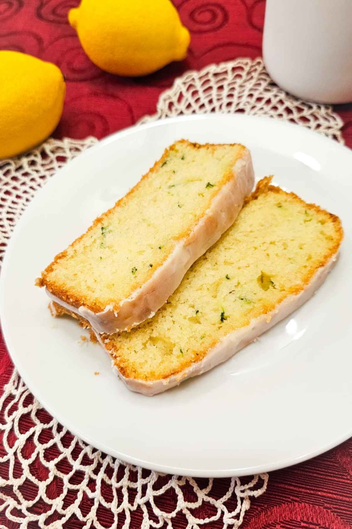 lemon zucchini bread on a white plate with a doily under it with a lemon to the side of it on a red vintage table cloth