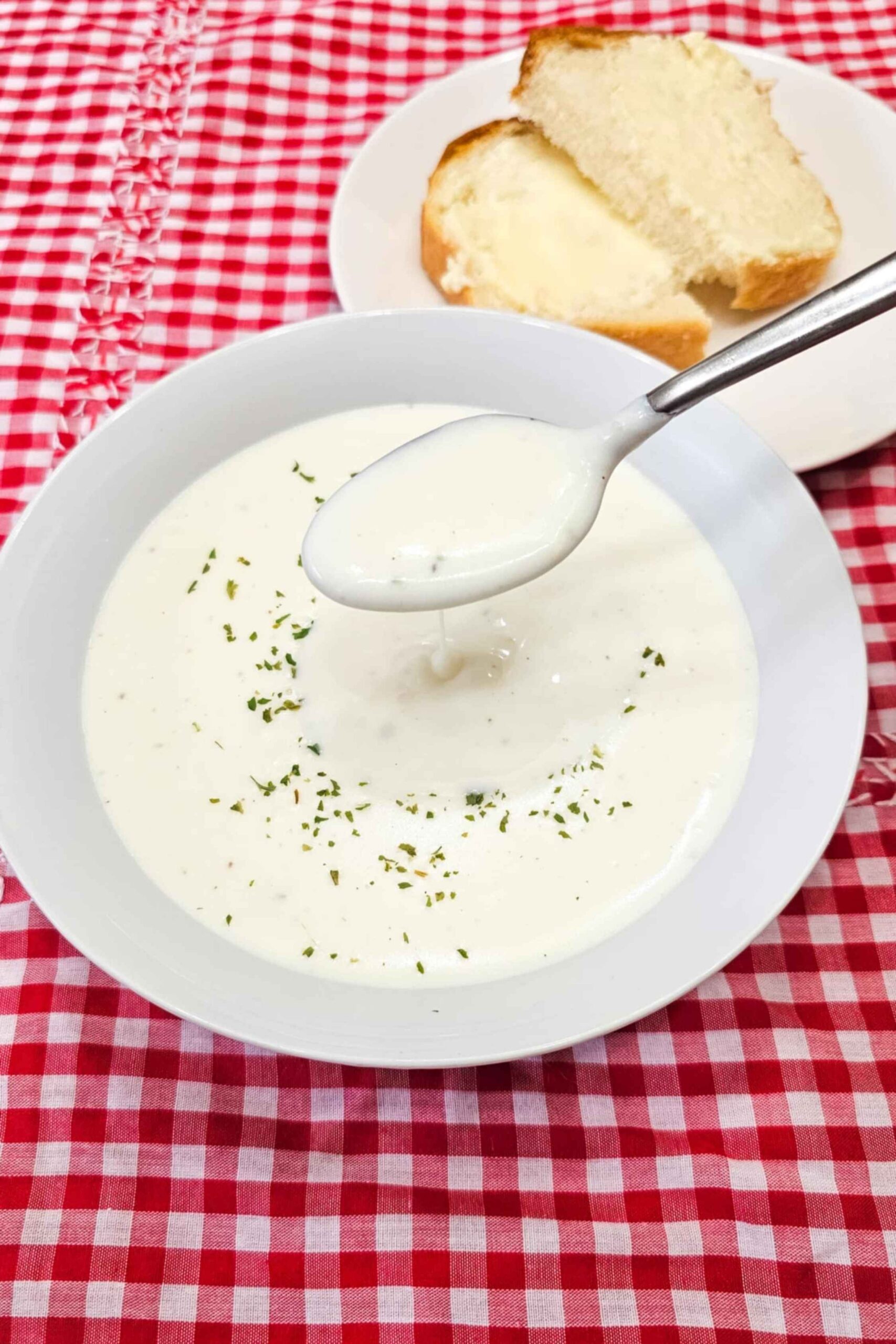 milk soup in a glass white bowl being spooned out, garnished with parsley and sitting on a vintage checkered red and white table cloth