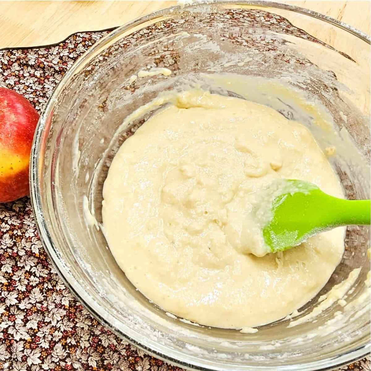 apple cake batter in a glass bowl with a silicon spatula in it with a fall cloth napkin to the side of it