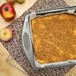 apple cake in a pan sitting on a wire rack on a fall cloth napkin sitting on a wooden counter top with fresh apples to the side