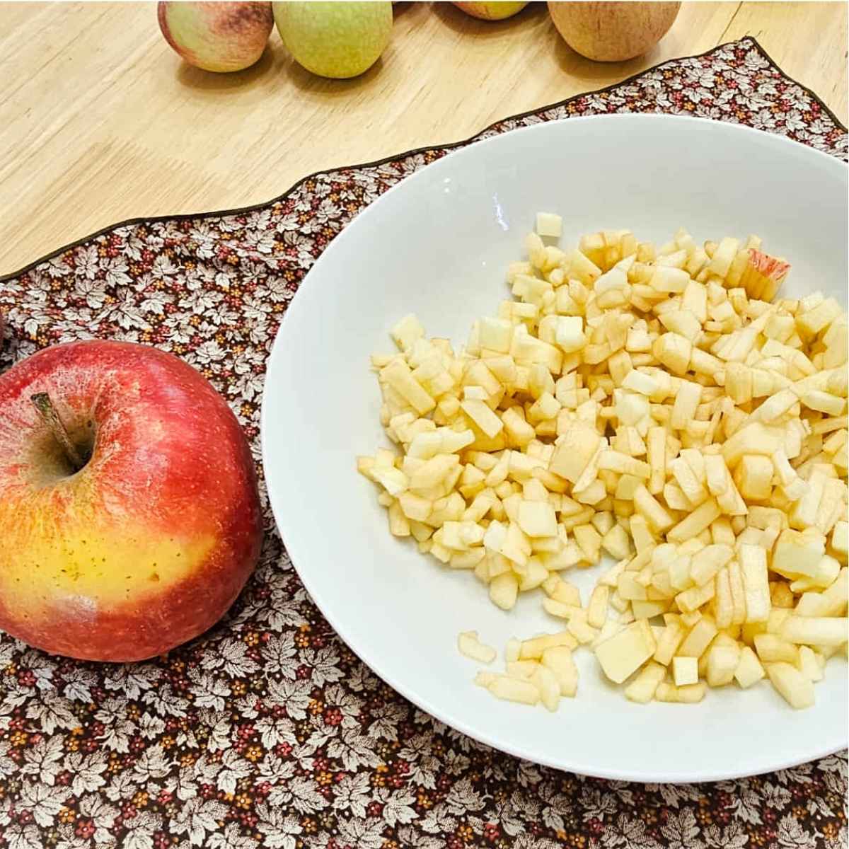 finely chopped apple in a white glass bowl sitting on a fall cloth napkin with fresh apples to the side