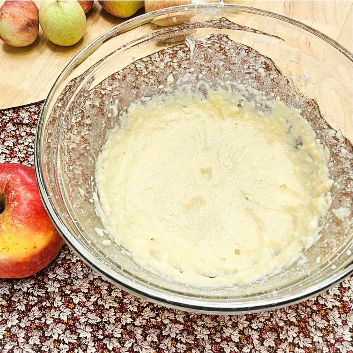 butter and sugar creamed together in a clear glass bowl and mixed with an egg and vanilla sitting on a fall cloth napkin with apples to the side of it