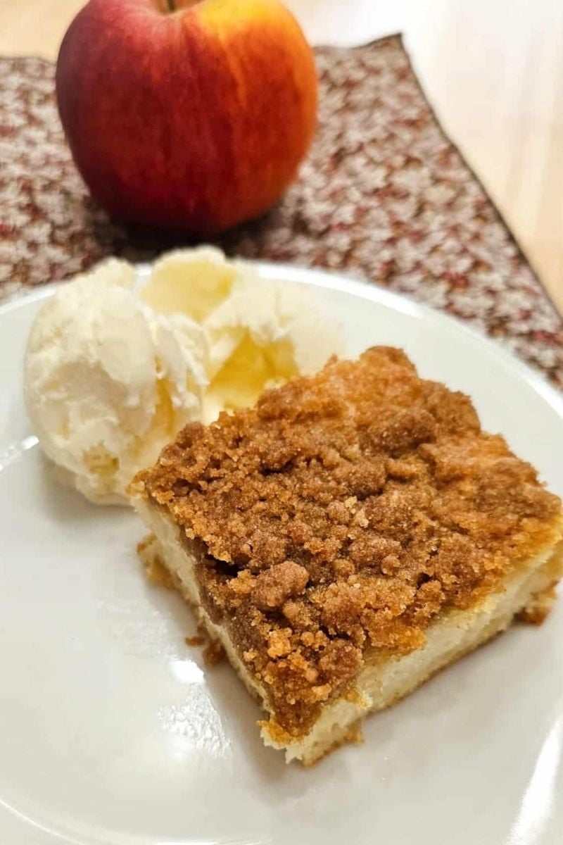 apple cake on a white plate with a scoop of vanilla ice cream to the side of it with a fresh apple and a fall cloth napkin on a wooden counter top