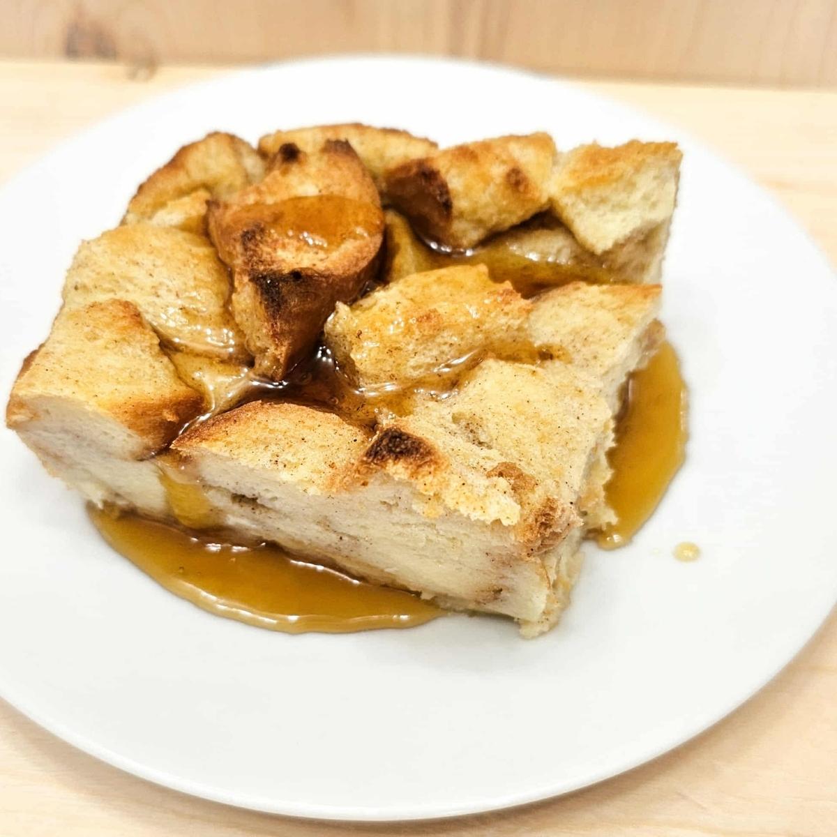 bread pudding topped with pure maple syrup and sitting on a white plate on a wooden counter top