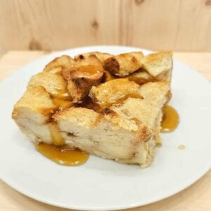 bread pudding sitting on a white plate and drizzled with pure maple syrup on a wooden counter top with a wooden background