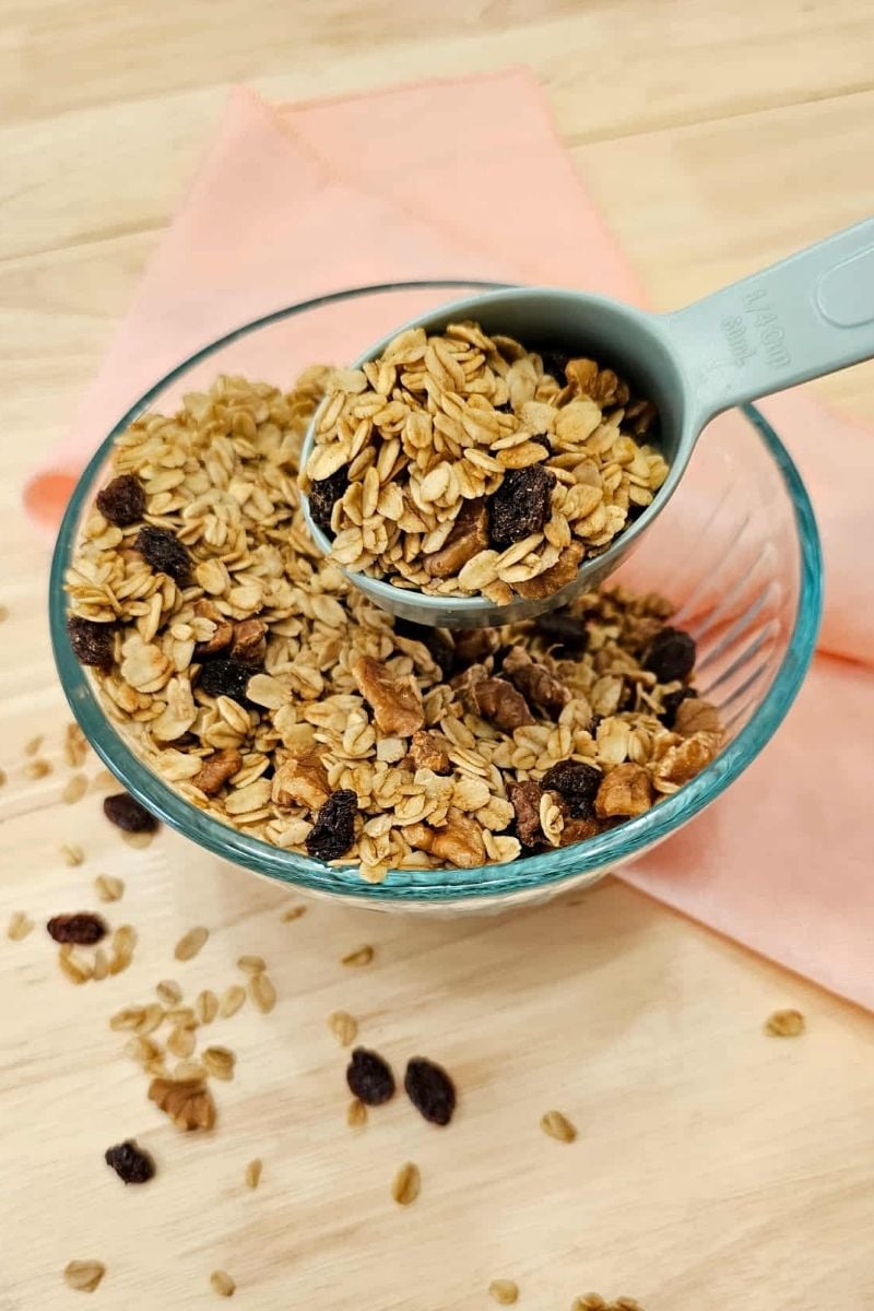 maple walnut granola sitting in a glass bowl on a wooden counter top with a pink cloth napkin under the bowl with a measuring spoon in it
