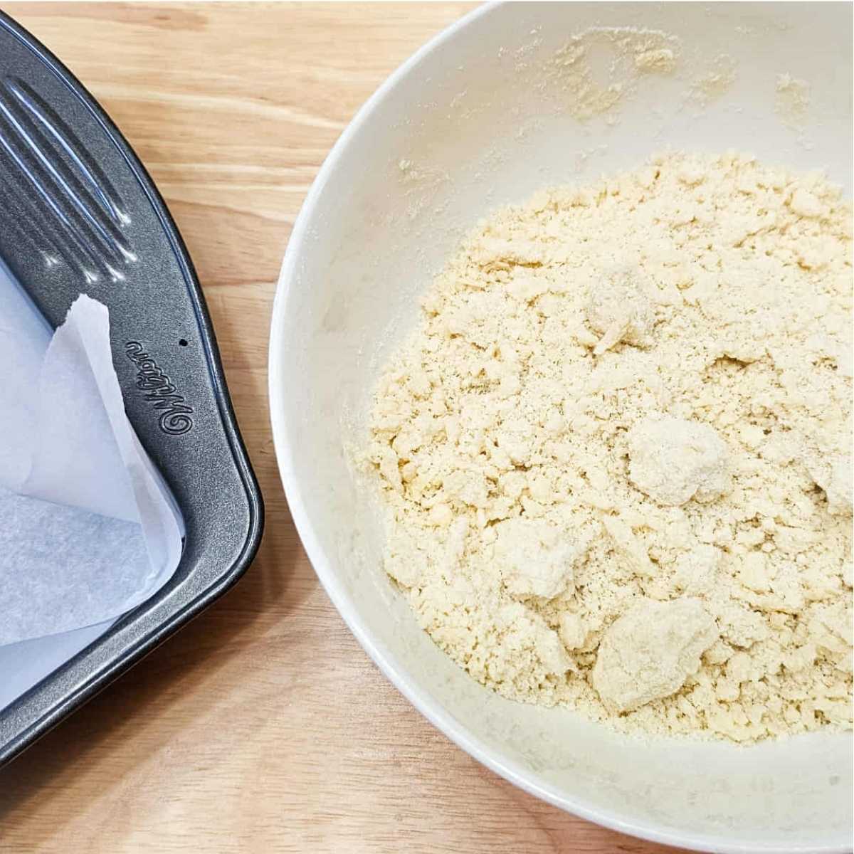 icing sugar, butter and flour in a white glass bowl with a parchment lined 9x9 pan next to it on a wooden counter top