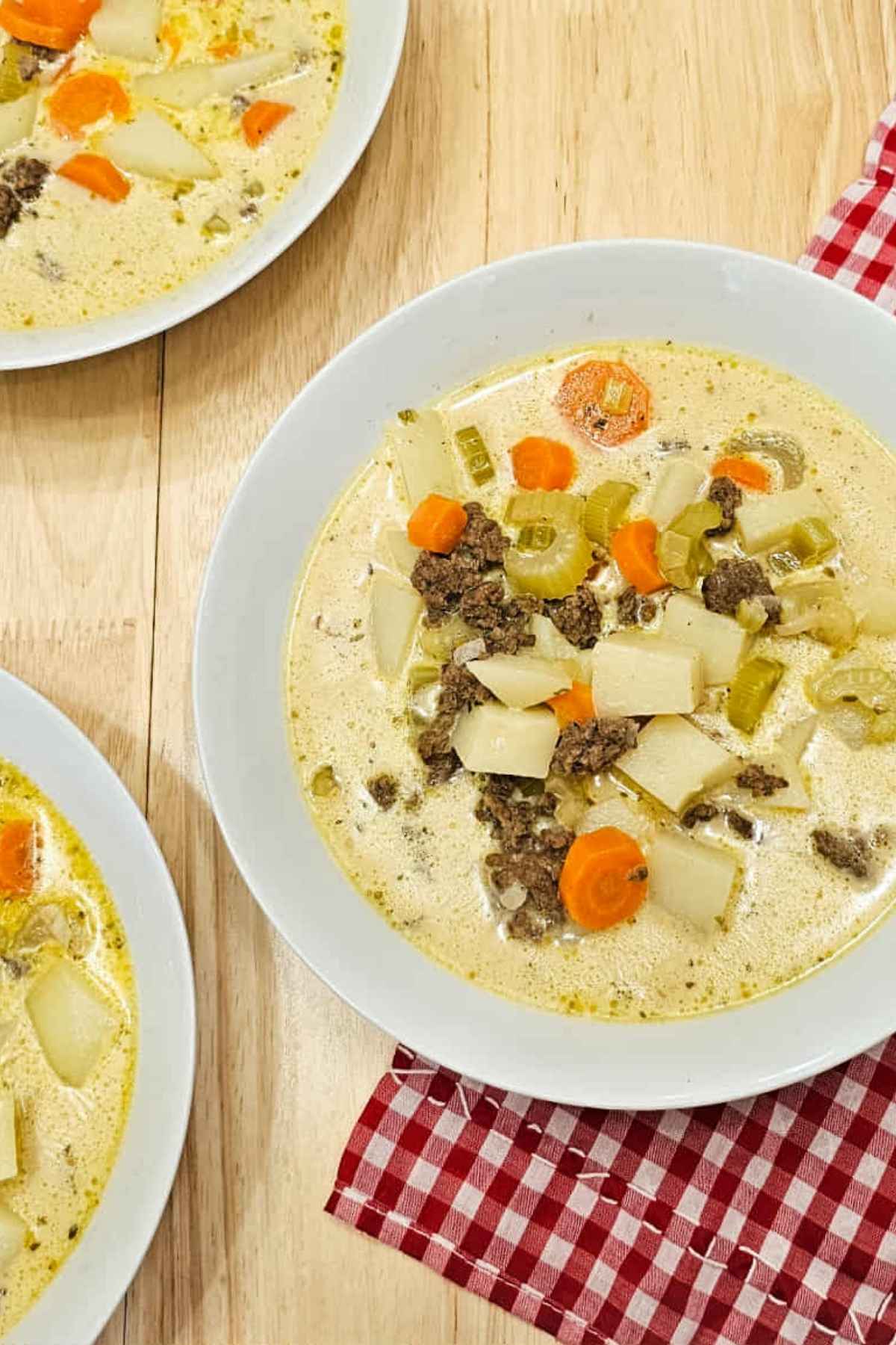 cheeseburger soup in 3 white glass bowls on a wooden counter top with a red checkered cloth napkin under a bowl