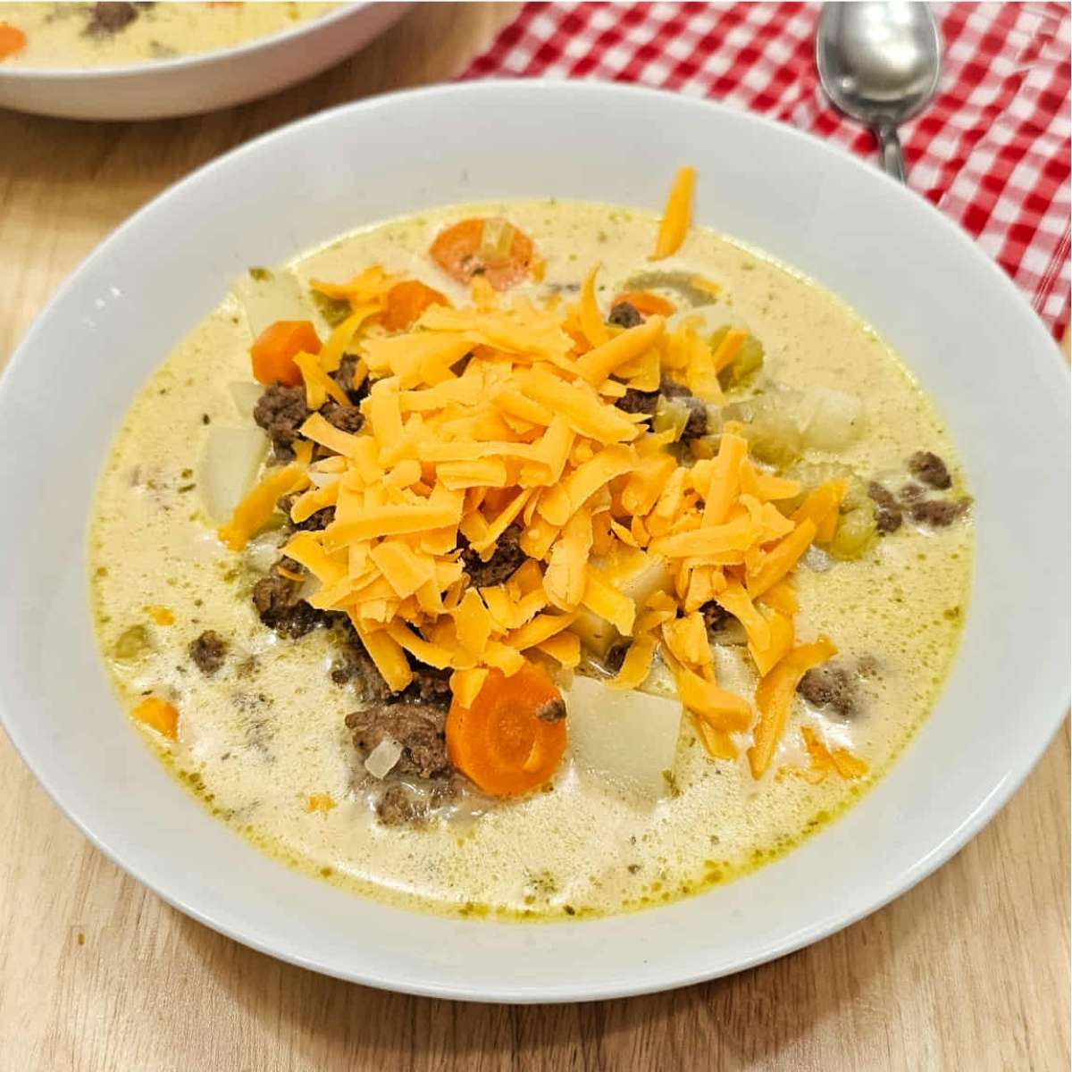 cheeseburger soup topped with shredded cheese sitting on a wooden counter top with a white and red checkered cloth napkin with a spoon on it