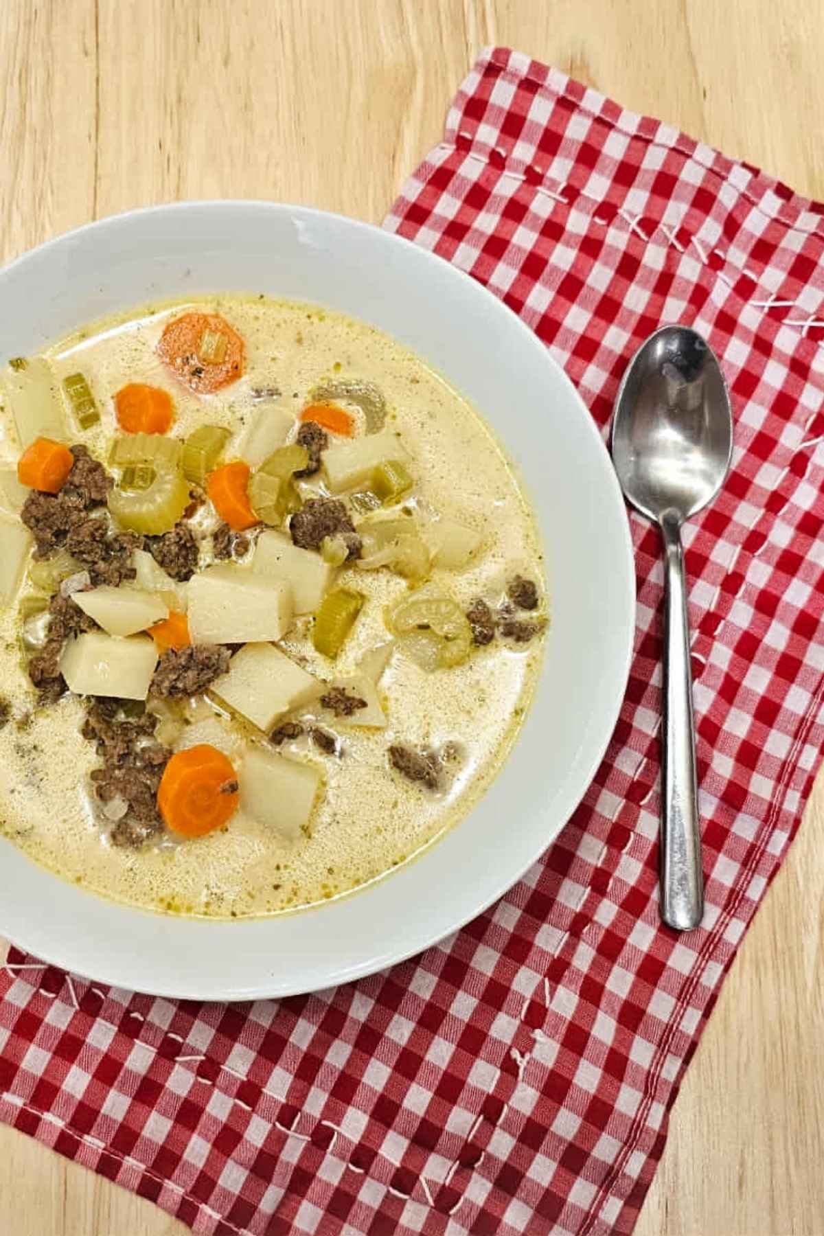 cheeseburger soup in a white bowl with a spoon next to it thats sitting on a red and white checkered cloth napkin on a wooden table