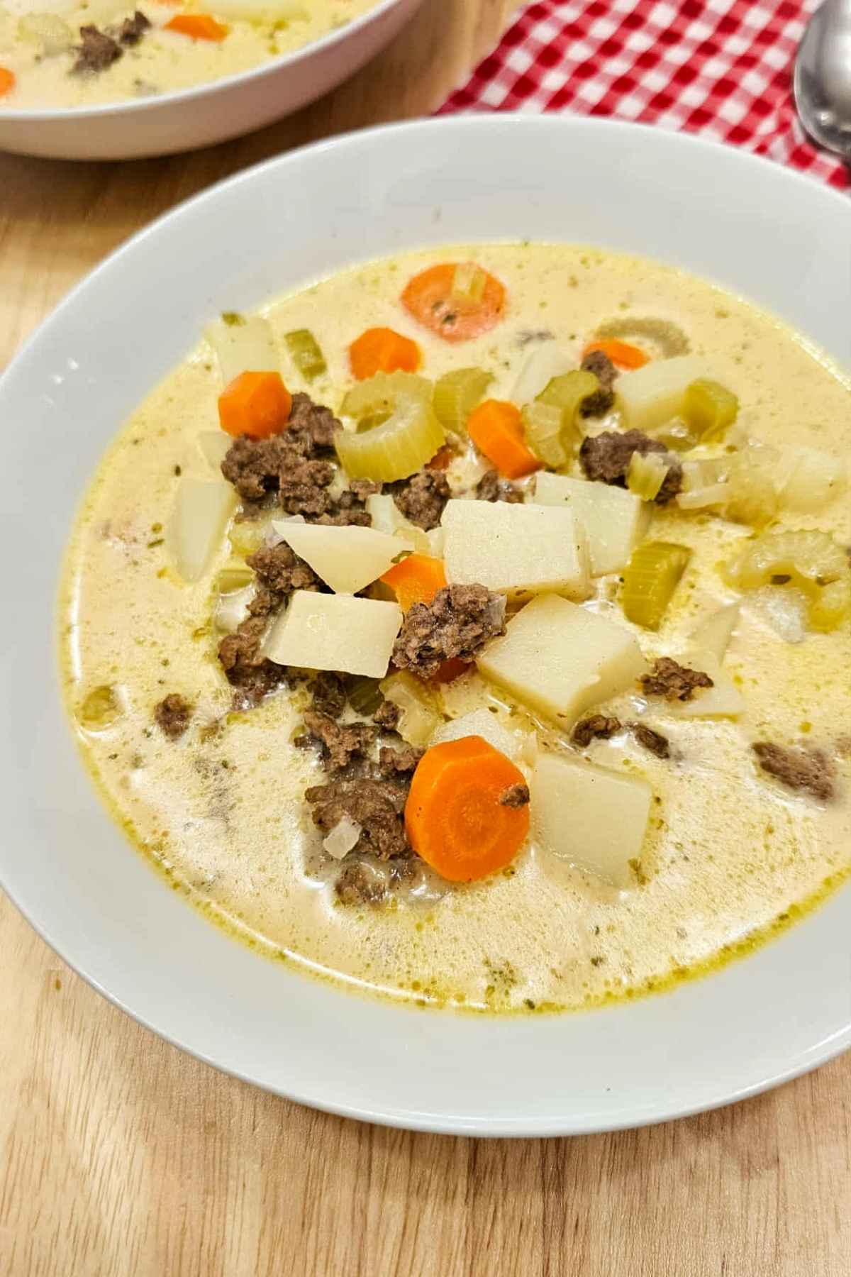 cheeseburger soup in a white glass bowl sitting on a wooden counter top with a close up sot of it, loaded with potatoes, ground beef, celery, carrots, onions and cheese