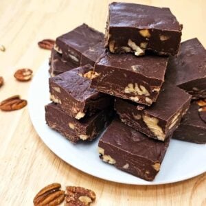 chocolate pecan fudge sitting on a white glass plate on a wooden counter top with pecans all around