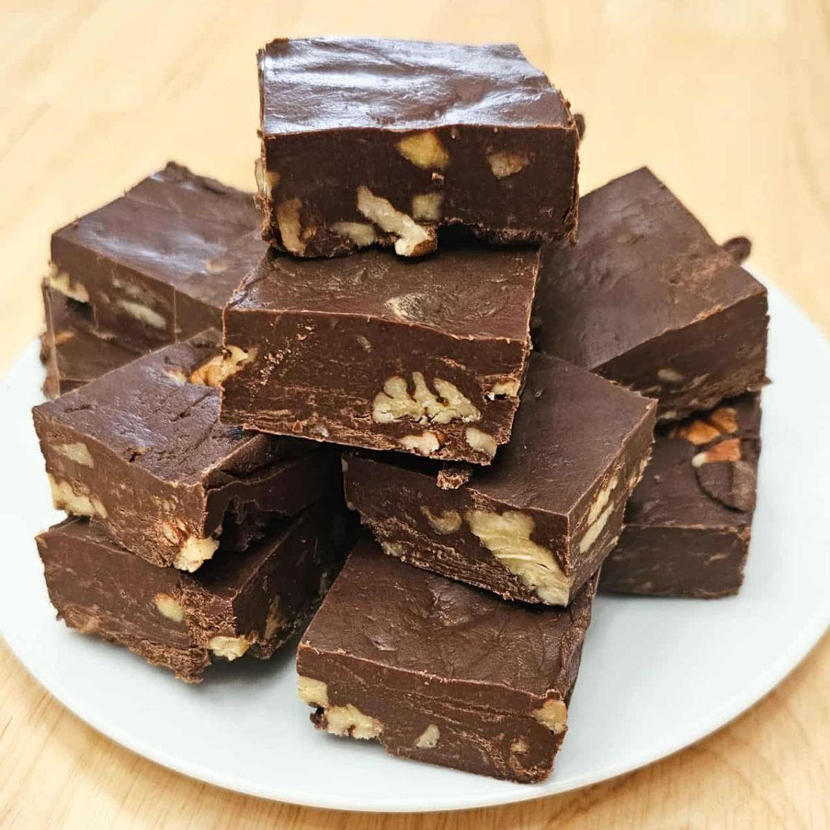 chocolate pecan fudge sitting in a pile on a white plate on a wooden counter top