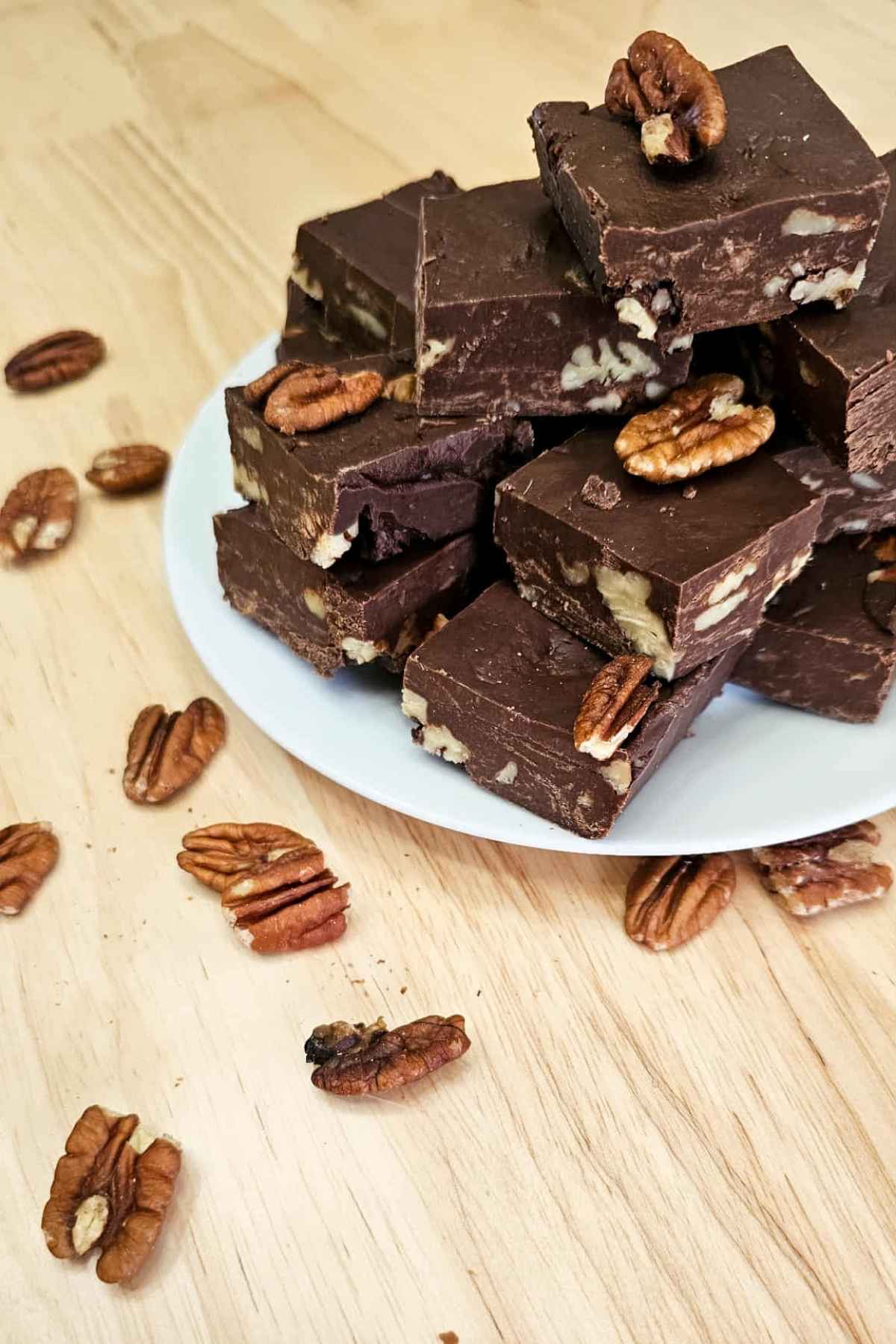 chocolate pecan fudge sitting on a white plate in a pile on a wooden counter top and surrounded by pecans