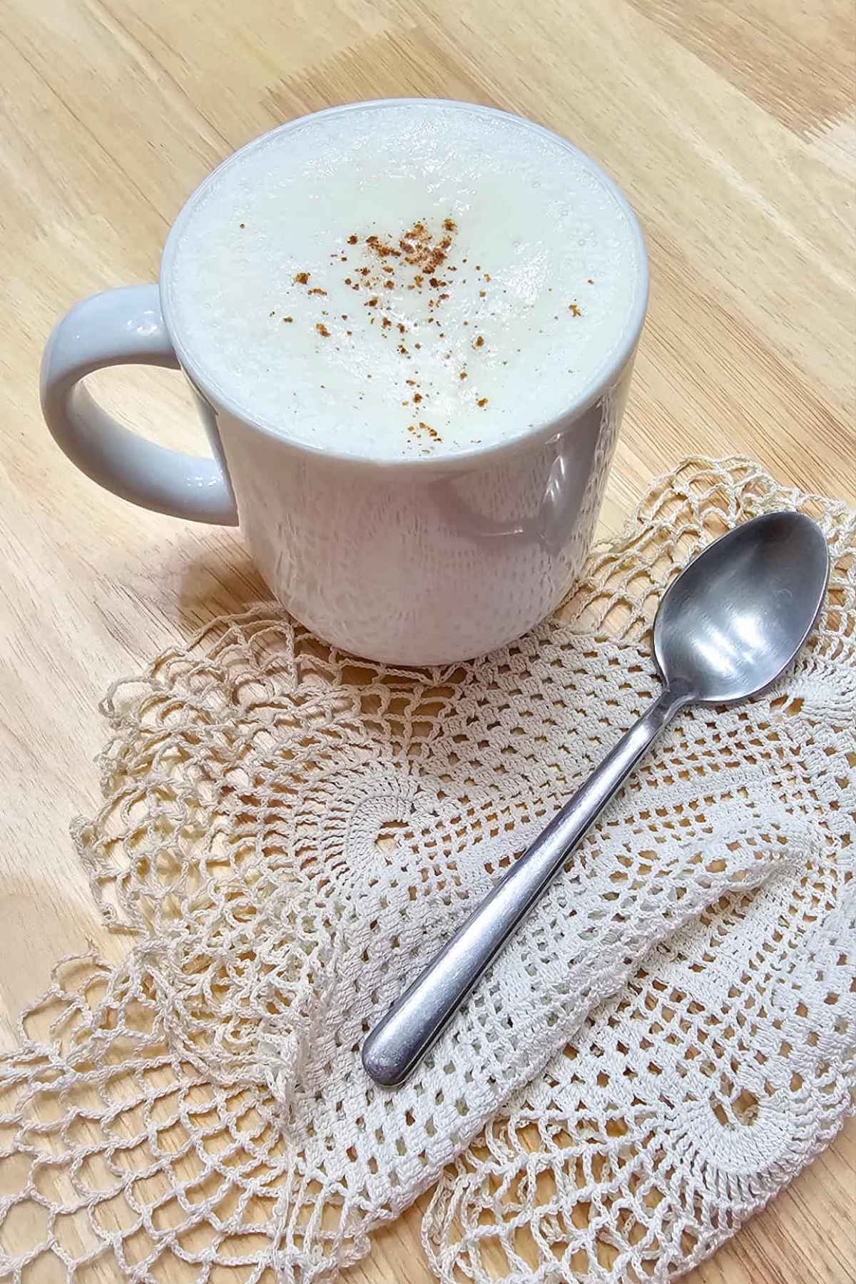 a glass of hot maple milk topped with homemade whip cream and a dash of cinnamon sitting on a doily with a spoon to the side on a wooden table