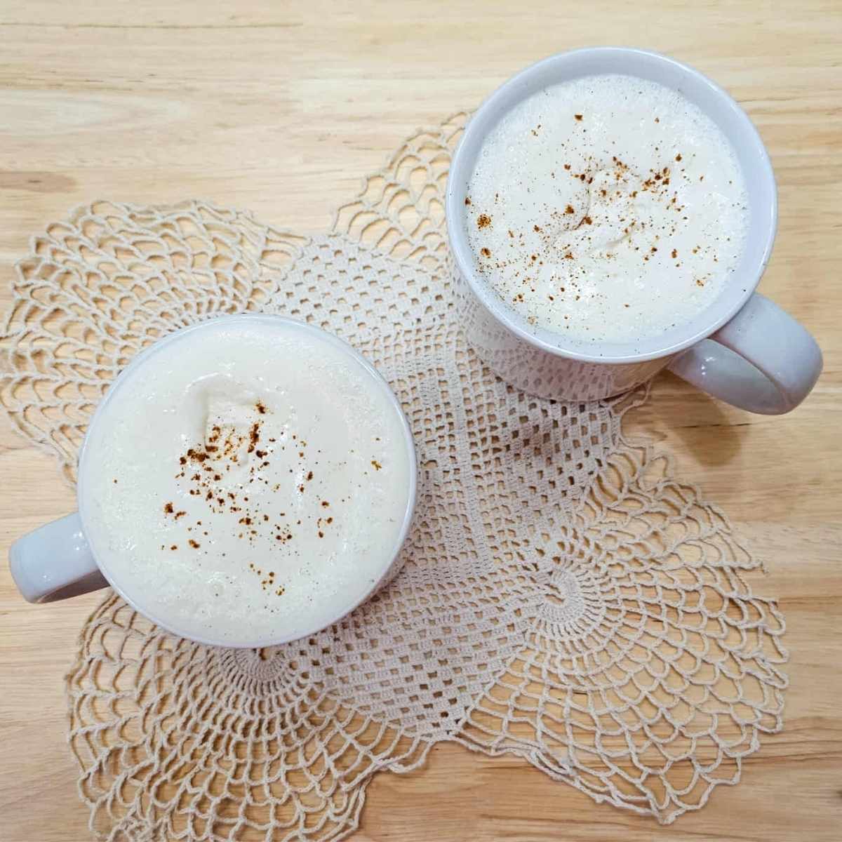 hot maple milk in 2 glasses sitting on a doily on a wooden counter top and topped with homemade maple syrup whip cream and a dash of cinnamon