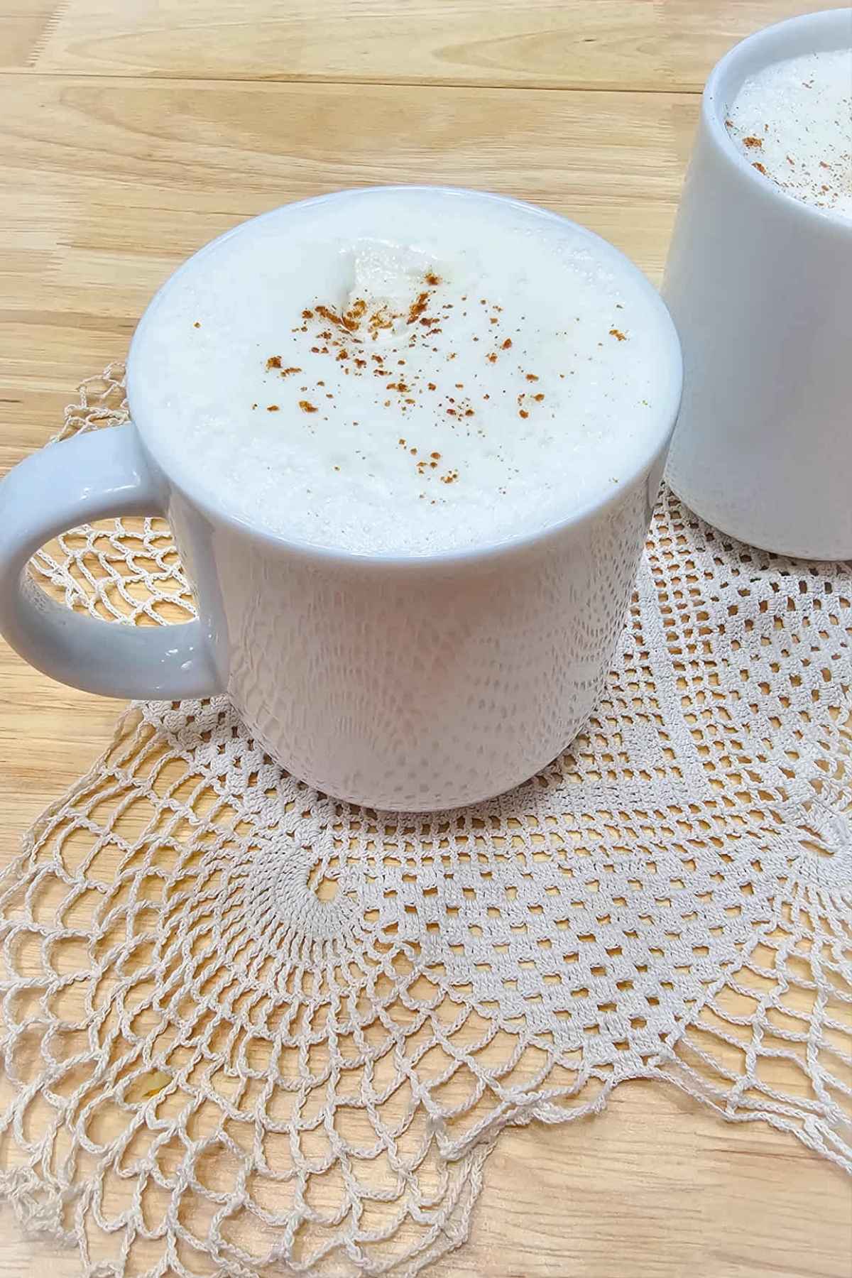 hot maple milk sitting on a doily on a wooden counter top and topped with pure maple syrup whip cream and a dash of cinnamon