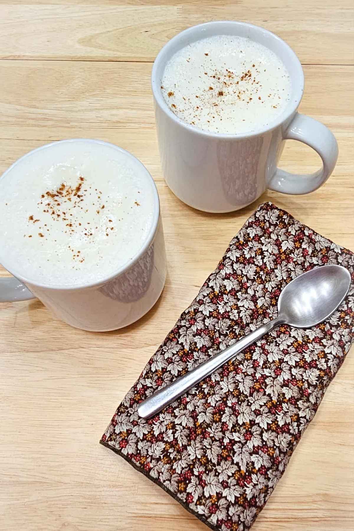 2 glasses of hot maple milk in white mugs, topped with homemade maple whip cream and a dash of cinnamon with a fall cloth napkin next to them with a spoon on top on a wooden table