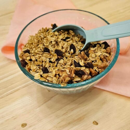 maple walnut granola sitting in a clear glass bowl with a measuring cup sitting in it on a wooden counter top with a pink cloth napkin under the bowl