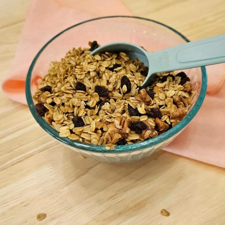 maple walnut granola sitting in a clear glass bowl with a measuring cup sitting in it on a wooden counter top with a pink cloth napkin under the bowl