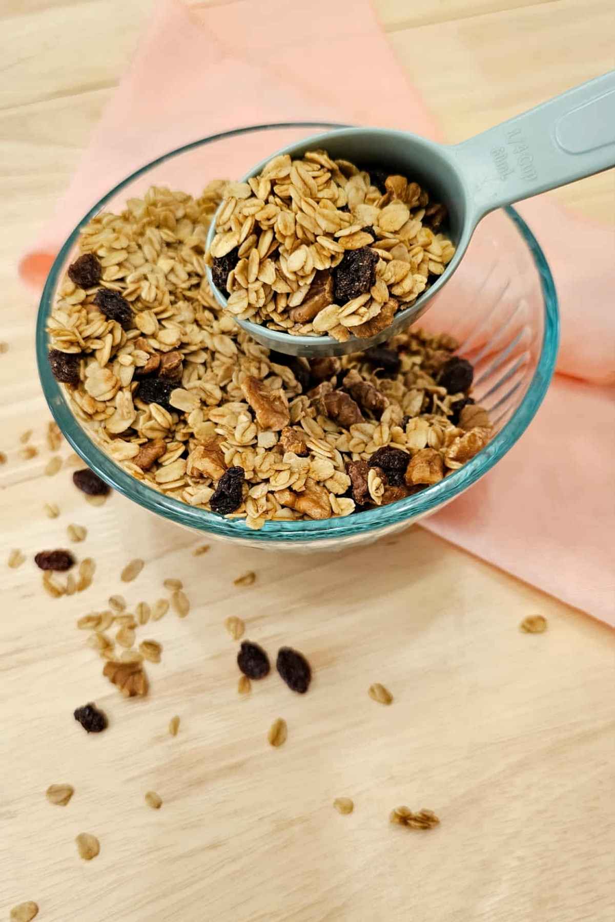 maple walnut granola sitting in a glass clear bowl being scooped out with 1/4 cup measuring cup and sitting on a wooden counter top with a pink cloth napkin to the side