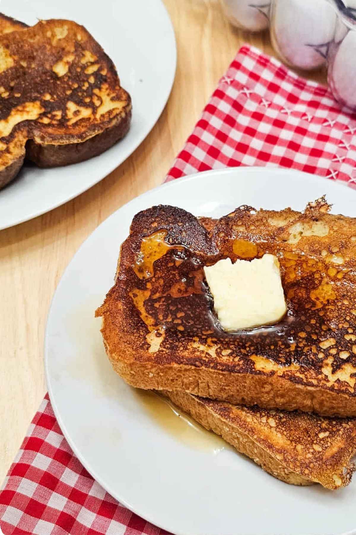 French toast sitting on a white glass plate on a red and white checkered cloth napkin with fresh eggs to the side of it and another plate with French toast