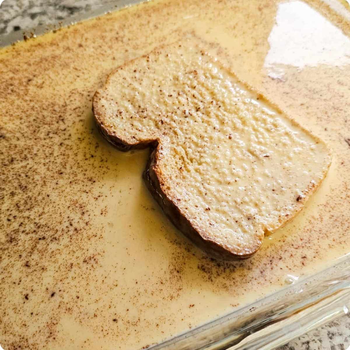 French toast being dipped into the custardy mixture in a 9x13 shallow glass dish sitting on a white and grey counter top