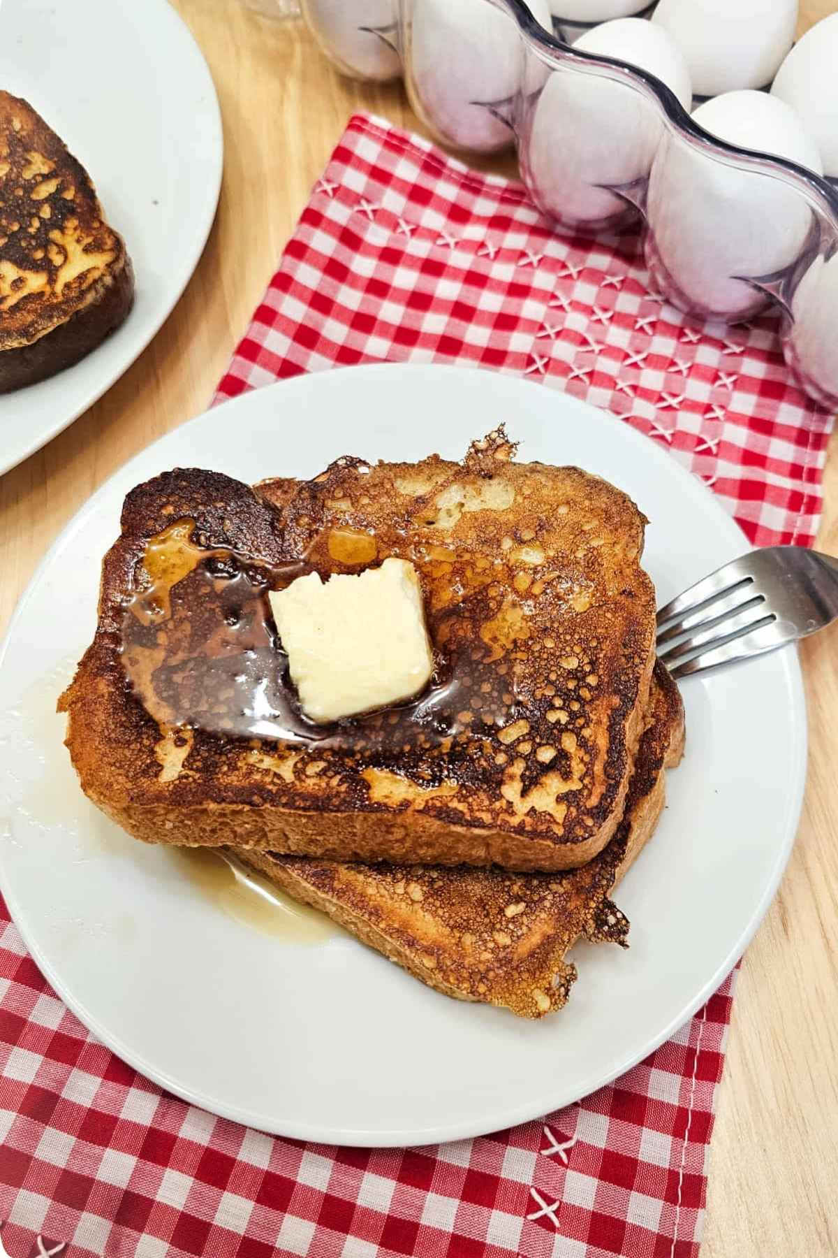 French toast sitting on a white plate with a dollop of butter and pure maple syrup drizzled over it with a fork on the plate next to a red and white checkered vintage cloth napkin