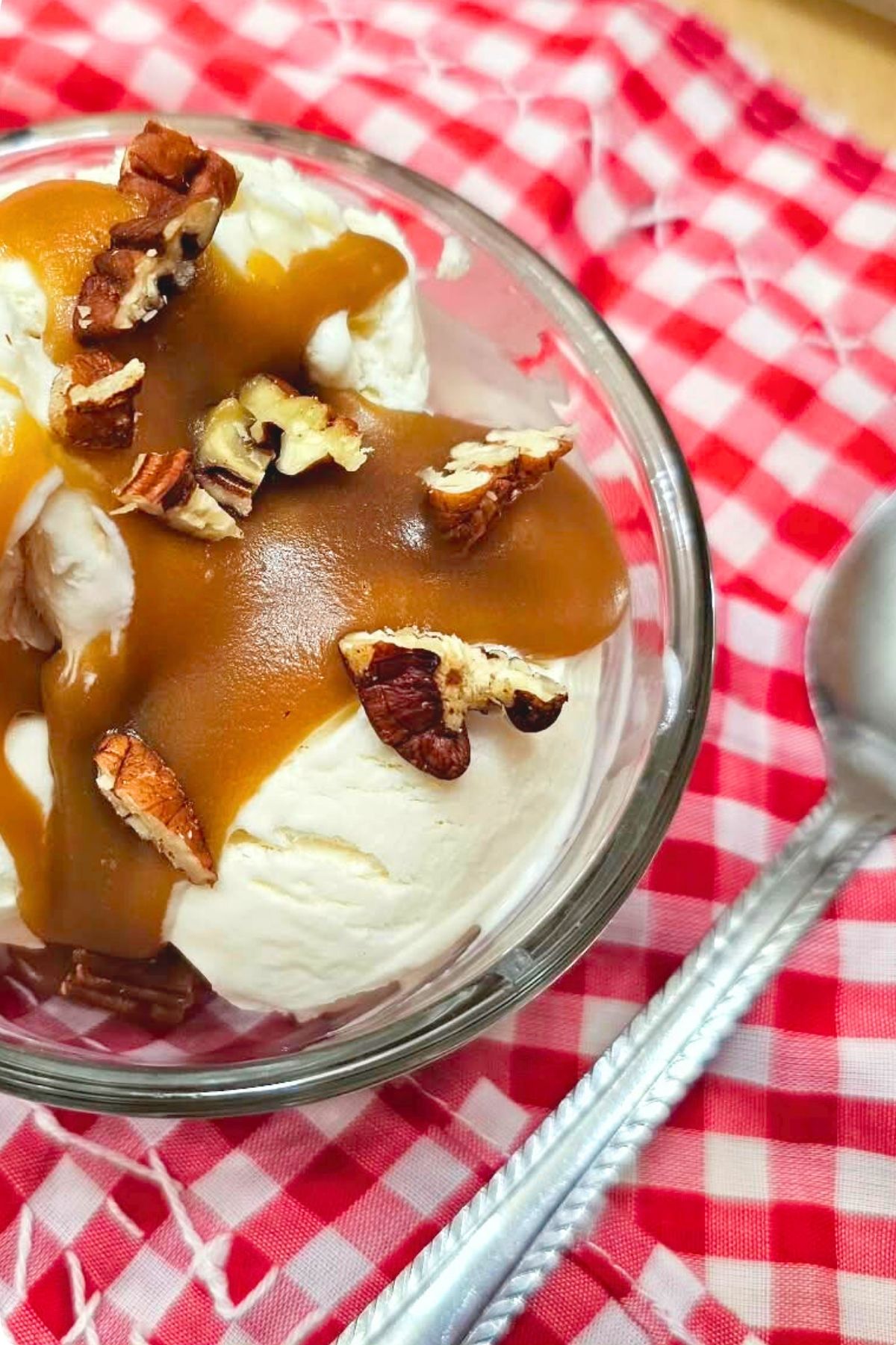Butterscotch sauce poured over homemade vanilla ice cream and topped with chopped pecans sitting on a red and white table cloth with a spoon to the side of it