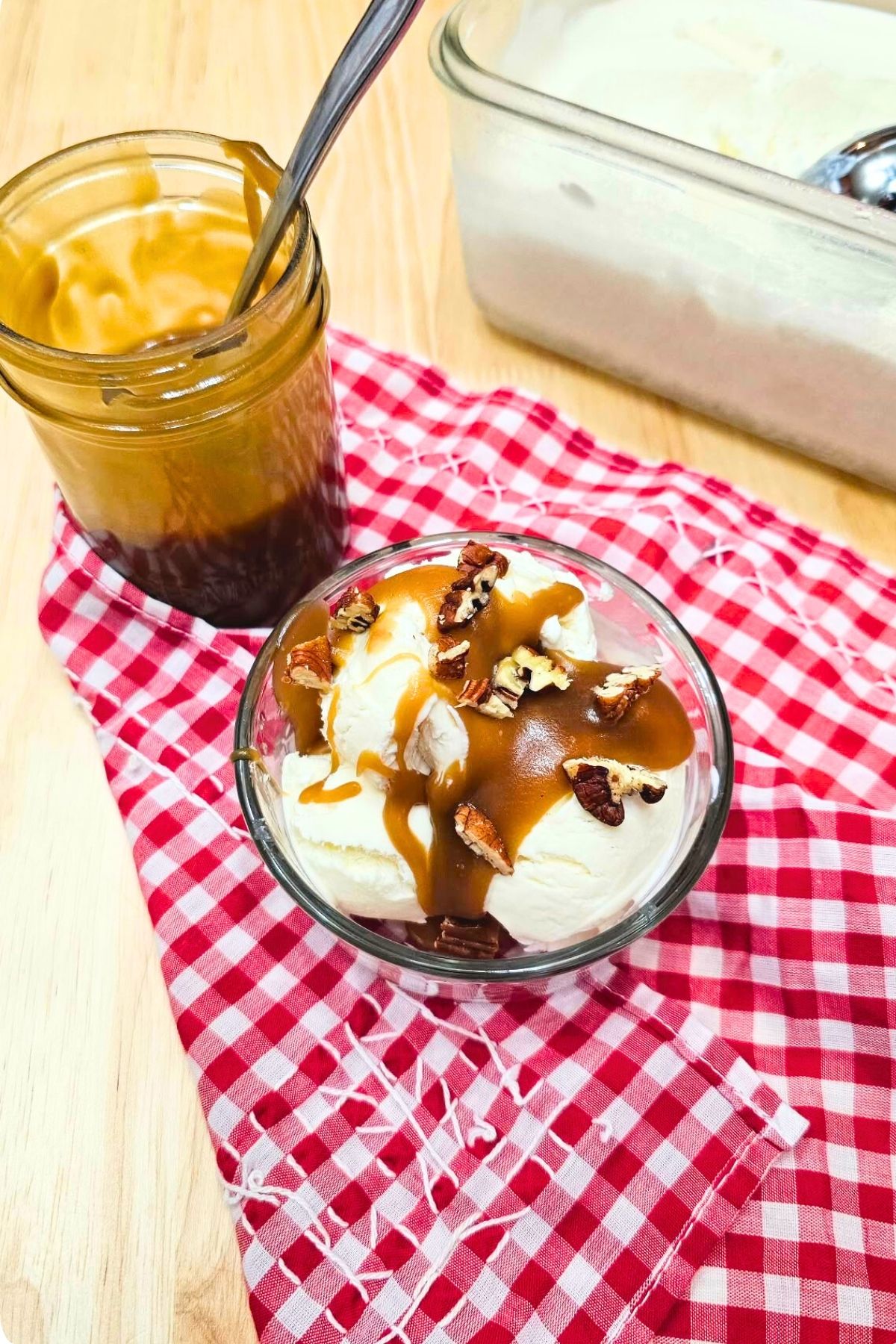 butterscotch sauce sitting in a mason jar on a red and white cloth napkin with a bowl of ice cream next to it with butterscotch sauce over it and diced pecans